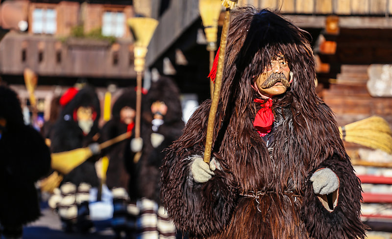 Carnival parade with bear-like man in the foreground wearing a fur coat with a hood and a mask.