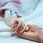 Close up on the holding hands of a mother and her recovering child, lying in a hospital bed. 