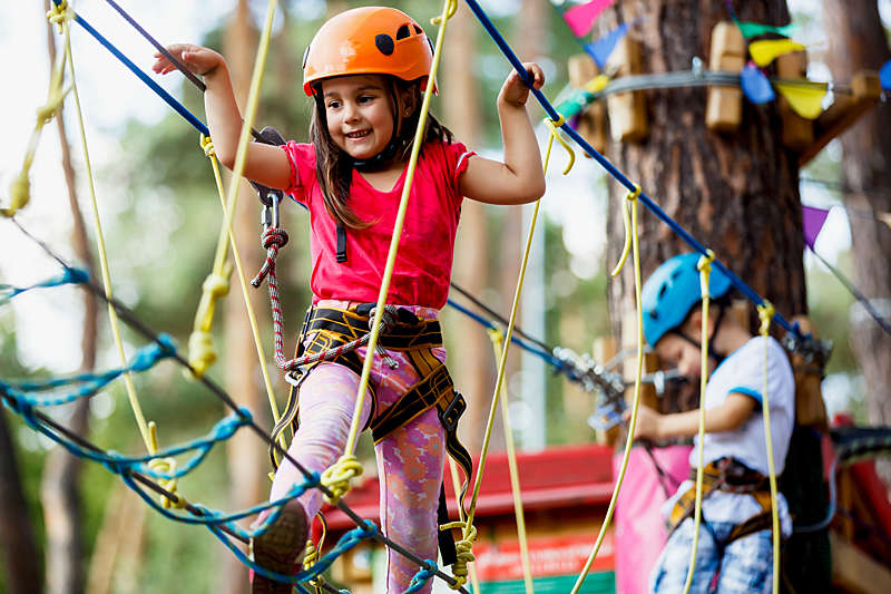 Little beautiful girl climbs on rope harness in a summer city park.