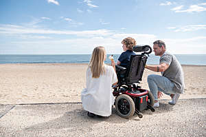 Parents kneeling by a 6 year old boy in a wheelchair, enjoying an ice cream on the beach, while looking at the view.