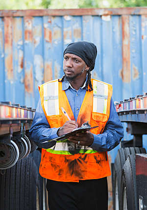 A man wearing a safety vest and taking notes, is standing between two semi trucks.