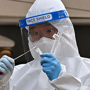 Close-up of a medical staff member in full protective gear holding a nasal swab and test tube.