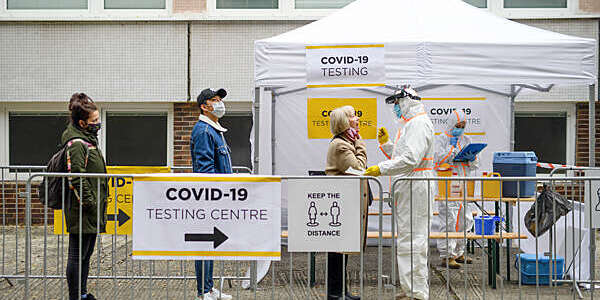 Three people line up on the street at a COVID-19 testing centre where a medical staff member in full white protective overalls performs the PCR swab test.