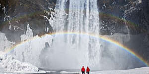 View of a couple in red anoraks dwarfed by a large waterfall with a rainbow shimmering through it, in Skogafoss, Iceland.