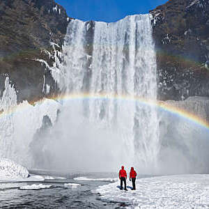 Couple under a rainbow by a large waterfall, in winter in Skogafoss, Iceland.